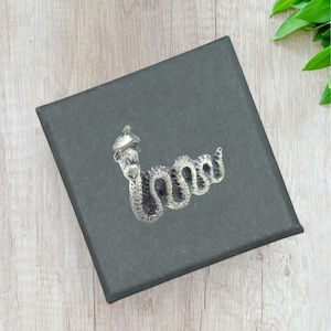May include: A silver-toned snake brooch with a hat, resting on a dark gray box. The snake is detailed with scales and a smiling face. The box is on a light wood surface, with green leaves in the corner.