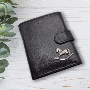 May include: Black leather wallet with a textured finish and a silver rocking horse emblem. The wallet has a flap closure and a button clasp. The background is a white wooden surface with green foliage.