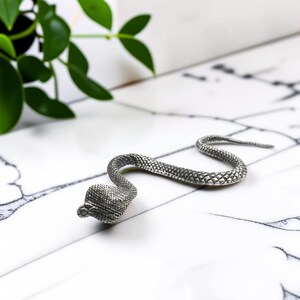 May include: A silver-toned metal snake figurine featuring a textured, scaled surface. The snake is depicted in a curved, S-shaped position with its head slightly raised, showcasing detailed markings across its entire body and tail.