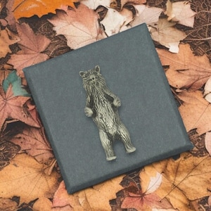 May include: A silver-coloured bear figurine standing upright on a dark grey box. The bear has detailed fur and is set against a backdrop of autumn leaves in shades of brown, orange, and yellow.