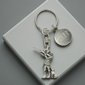 May include: Silver-coloured keyring featuring a hunter aiming a rifle, with a dog at their side. The keyring includes a round tag with the initials "MH" engraved. The metal has a slightly aged appearance and the design is detailed.