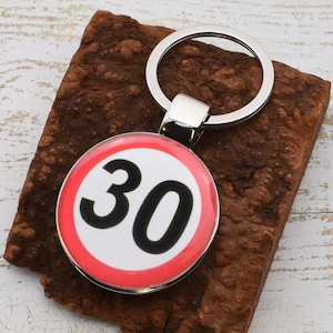 May include: A silver metal keyring with a circular charm displaying a speed limit sign. The sign shows the number 30 in black on a white background, with a red border. The keyring rests on a textured, brown surface.