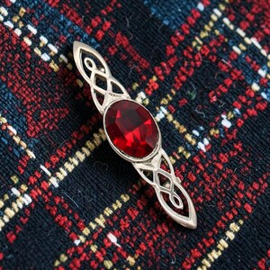May include: A silver-toned metal brooch featuring an elongated, pointed design with intricate openwork knot patterns on both ends. A large, oval-shaped, deep red faceted gemstone is securely set in the centre of the piece, creating a striking contrast.