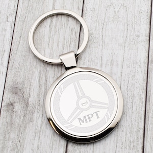 Op de afbeelding: Een zilverkleurige metalen sleutelhanger met een ronde hanger. De hanger heeft een stuurwielontwerp met de letters "MPT" en een hoornsymbool. De sleutelhanger en hanger zijn verbonden door een klein, rechthoekig stuk.