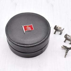 May include: A black leather cufflink box with a red and white flag design on the lid. The box is open and shows two silver cufflinks with a goat design and one silver cufflink with a caduceus design.