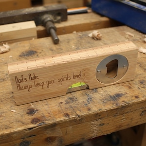 Puede incluir: Un nivel de madera con un abrebotellas y el texto grabado "Dad's Rule: Always keep your spirits level." El nivel tiene un abrebotellas de metal y un nivel de burbuja. La madera tiene marcas de regla a lo largo del borde superior.