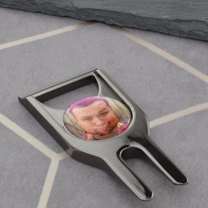 Könnte beinhalten: Ein schwarzer Golfball-Retriever aus Metall mit einem runden, transparenten Kunststoffeinsatz, der ein Foto einer Person mit pinkfarbenem Haar zeigt.