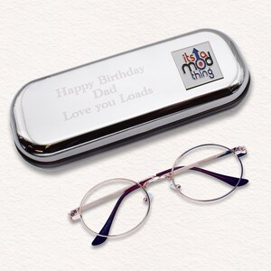 May include: A silver eyeglass case with the inscription "Happy Birthday Dad Love you Loads" and a small logo that says "its a mod thing".  A pair of round, gold-rimmed reading glasses are lying next to the case.