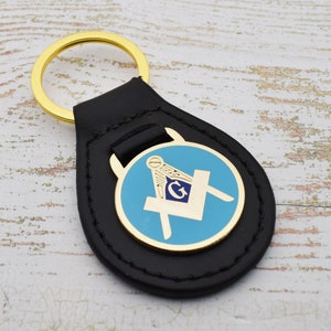 Op de afbeelding: Een zwarte leren sleutelhanger met een gouden metalen hanger met een blauw en gouden vrijmetselaars symbool.