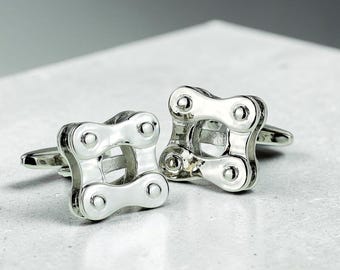 May include: A pair of silver-toned cufflinks shaped like square segments of a bicycle chain. Each cufflink features a polished metallic finish with four circular rivet details at the corners and a hollow center, designed with a standard toggle backing.