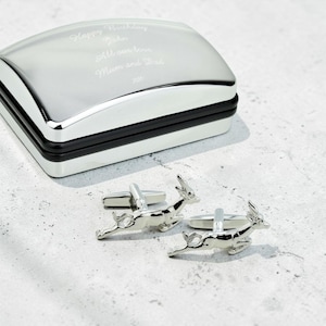 May include: A pair of silver-toned metal cufflinks shaped like leaping deer, positioned in front of a rectangular silver-toned presentation box with a black trim. The lid of the box is engraved with the text: Happy Birthday John All our love Mum and Dad xxx.