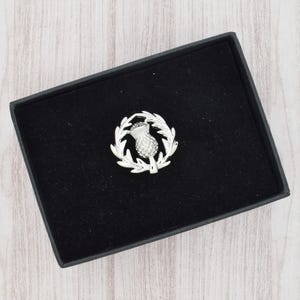 Pode incluir: Um broche de cardo prateado, apresentado numa caixa preta. O broche apresenta um design detalhado de cardo rodeado por uma coroa de folhas. A caixa é retangular e tem um interior de veludo preto.