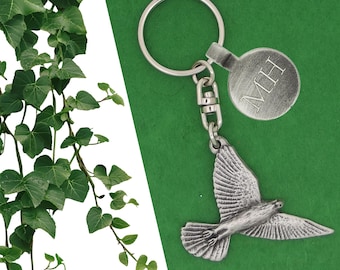Puede incluir: Un llavero plateado con un colgante detallado de un pájaro volando. El llavero incluye una etiqueta redonda con las iniciales "MH" grabadas. El fondo es de color verde liso, con hiedra verde en la esquina superior izquierda.