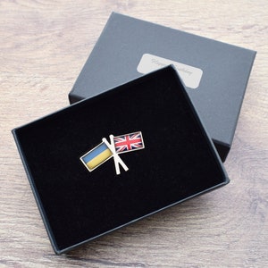 Puede incluir: Un pequeño pin de plata con banderas cruzadas de Ucrania y el Reino Unido. El pin está en una caja de terciopelo negro con las palabras "Feliz cumpleaños" en la tapa.