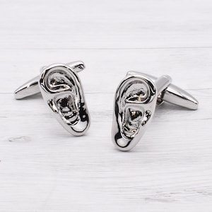 May include: A pair of silver cufflinks shaped like human ears. The cufflinks are detailed and realistic.