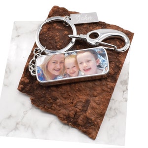 Könnte beinhalten: Silberner Schlüsselanhänger mit einem rechteckigen Fotoeinsatz, der eine dreiköpfige Familie zeigt. Das Foto zeigt eine Frau und zwei Kinder, die lächeln. Der Schlüsselanhänger hat außerdem einen silbernen Ring und einen silbernen Haken. Das Schild am Schlüsselanhänger lautet "John".