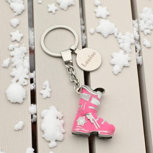 Puede incluir: Un llavero plateado con un dije de bota de esquí rosa y una etiqueta redonda grabada con el nombre "Louise". La bota de esquí tiene detalles blancos y un diseño de estrella. El llavero está sobre una superficie clara con decoraciones de copos de nieve.