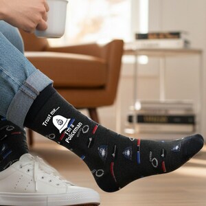 May include: Dark grey socks patterned with police-related motifs, including handcuffs and badges. The socks display the text "Trust me... I'm a Policeman". The socks are worn with blue jeans and white trainers.