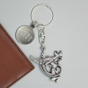May include: A silver-toned keyring featuring a dragon pendant and a circular tag engraved with "MH". The dragon has wings and a curled tail. The keyring is attached to a silver key ring.