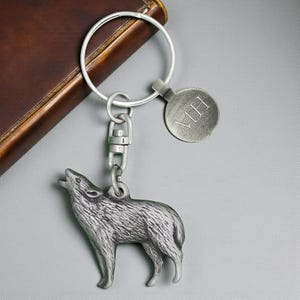 May include: A silver-colored keychain featuring a howling wolf pendant. The keychain includes a round tag with the initials "MH" engraved on it. The keychain is attached to a silver ring and a clasp.