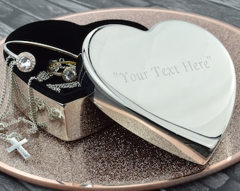 May include: A silver heart-shaped jewelry box with a black velvet interior. The box is open and shows a variety of jewelry, including a necklace with a cross pendant, a bracelet, and several rings. The box is engraved with the text "Your Text Here".