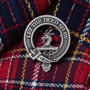 May include: A silver Shaw clan crest badge, featuring a stag's head within a belt inscribed with "LET THE DEED SHAW." The badge is set against a backdrop of red, blue, and white tartan fabric.