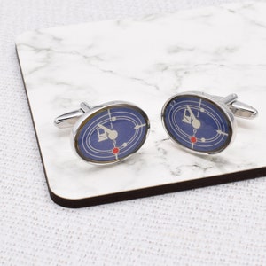 May include: A pair of silver cufflinks featuring a blue and white radar screen design. The screen has a red dot in the centre and a white arrow pointing to the right.