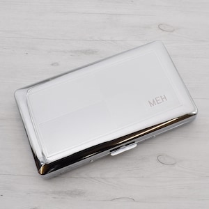 Op de afbeelding: Een zilveren sigarettenkoker met een getextureerd oppervlak en het woord "MEH" gedrukt op de voorkant.