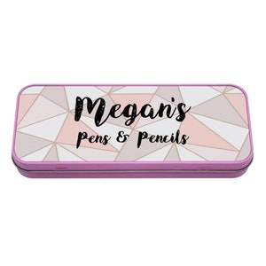 Puede incluir: Un estuche de lápices de metal rosa con un patrón geométrico en tonos rosa, blanco y dorado. El texto "Megan's pens & pencils" está impreso en el estuche.