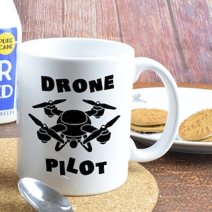 Puede incluir: Taza de cerámica blanca con una silueta negra de un dron y el texto "Drone Pilot".