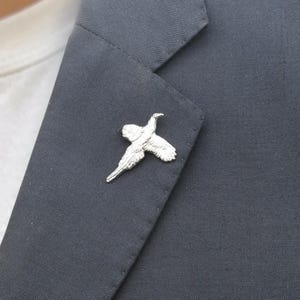 May include: A silver-toned metal lapel pin shaped like a bird in flight, featuring detailed wing and tail feather textures. The pin is attached to the dark charcoal gray fabric of a blazer, showcasing a clean, metallic finish and a sharp silhouette.