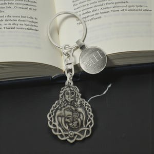 May include: A silver-toned keychain featuring a detailed pendant with a figure holding a bird, surrounded by Celtic knotwork. The keychain includes a round tag with the initials "MH". The background includes an open book.