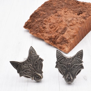 May include: Two silver wolf head cufflinks with intricate Celtic knot designs. The cufflinks are on a white surface.