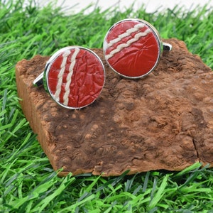 May include: A pair of silver cufflinks with red and white cricket ball designs. The cufflinks are on a piece of brown bark.