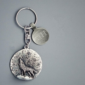 May include: A silver-coloured keyring featuring a howling wolf against a textured moon backdrop. Includes a small, round tag with the initials "MH". The keyring has a clasp and a key ring.
