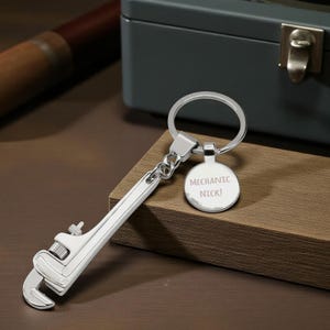 Puede incluir: Un llavero plateado con forma de llave inglesa y una etiqueta redonda con la inscripción "MECHANIC NICK!". El llavero de metal brillante descansa sobre una superficie de madera. En el fondo se ve una caja de herramientas gris.