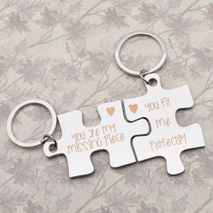 Puede incluir: Dos piezas de rompecabezas plateadas en forma de llaveros con el texto "You are my missing piece" y "You fit me perfectly" en letras doradas. Las piezas encajan para formar un corazón.