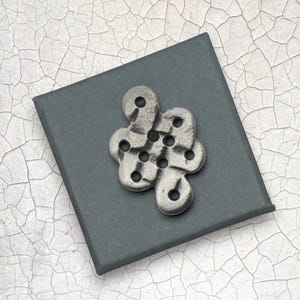 Può includere: Un ciondolo in metallo argentato con un design a forma di nodo, con molteplici aperture circolari. Il ciondolo è appoggiato su una scatola quadrata grigio scuro, su uno sfondo bianco e grigio screpolato.