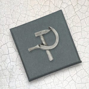 Puede incluir: Un emblema plateado de martillo y hoz, símbolo del comunismo, se muestra sobre un cuadrado gris oscuro. El emblema se coloca sobre una caja negra, sobre un fondo agrietado blanquecino.