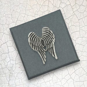 May include: A silver angel wing brooch on a dark grey box. The wings feature detailed, textured lines, and the box sits on a cracked white and grey surface. The brooch is a decorative accessory.