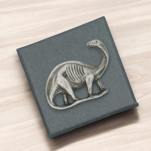 Könnte beinhalten: Ein silberfarbener Dinosaurier-Ansteckpin mit sichtbaren Rippendetails liegt auf einer dunkelgrauen Schachtel. Der Pin ist eine stilisierte Darstellung eines Brontosaurus. Die Schachtel ist quadratisch und hat eine strukturierte Oberfläche.