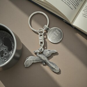 Pode incluir: Um porta-chaves prateado com um design detalhado de fénix com asas abertas. O porta-chaves inclui uma etiqueta redonda com as iniciais "MH" gravadas. O porta-chaves está preso a um fecho de metal.