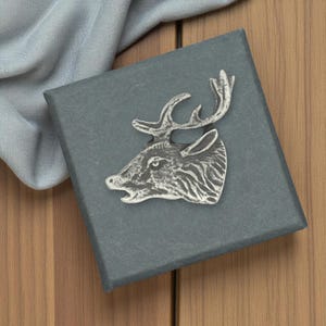 May include: A silver-coloured deer head brooch on a dark grey box. The brooch has detailed textured lines, antlers and an open mouth. A light blue fabric is draped over the box, set against a wooden background.