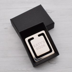 May include: A silver metal bookmark with the text "KATY'S BOOK" engraved on it, presented in a black gift box. The bookmark has a rectangular design with a curved top, perfect for marking pages in a book.