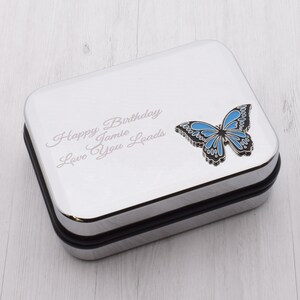 May include: A silver-colored rectangular box with a black border. The box features a blue and black butterfly design and the engraved text "Happy Birthday Jamie Love You Loads."