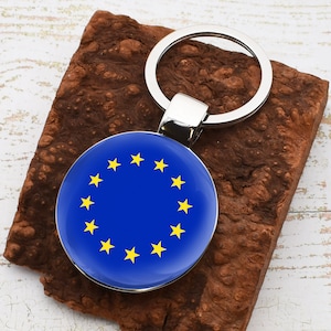 Puede incluir: Un llavero plateado con un charm redondo azul que presenta un círculo de estrellas amarillas. El charm es una representación de la bandera de la Unión Europea.