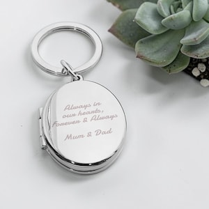 Könnte beinhalten: Ein silberner, ovaler Schlüsselanhänger mit graviertem Text: "Always in our hearts, Forever & Always Mum & Dad." Das Medaillon ist geöffnet und hat ein Scharnierdesign. Eine grüne Sukkulentenpflanze ist im Hintergrund zu sehen.
