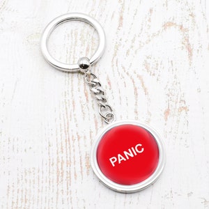Może przedstawiać: Srebrny brelok do kluczy z czerwonym, okrągłym przyciskiem, na którym widnieje napis "PANIC" białymi literami.