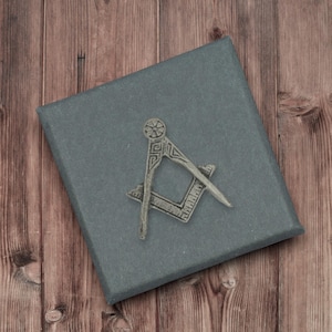 Puede incluir: Una caja gris con un símbolo masónico plateado, un diseño de compás y escuadra. La caja está sobre un fondo de madera.