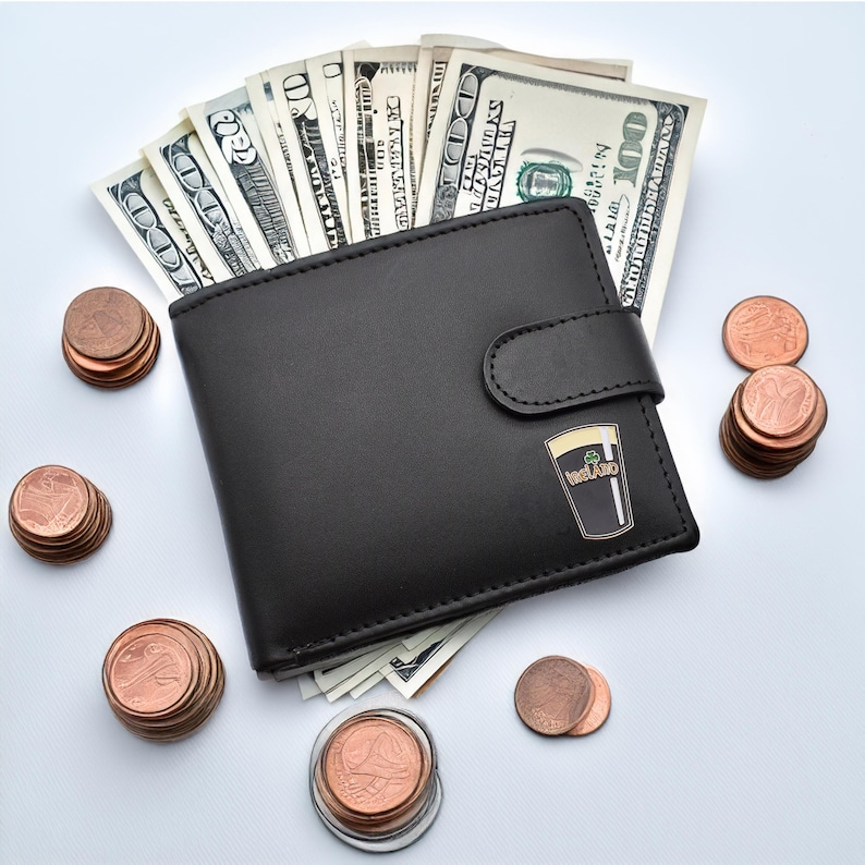 May include: A black leather wallet with a pin of a glass of Guinness beer with the word "Ireland" on it. The wallet is open and filled with US dollar bills. There are stacks of US pennies around the wallet.